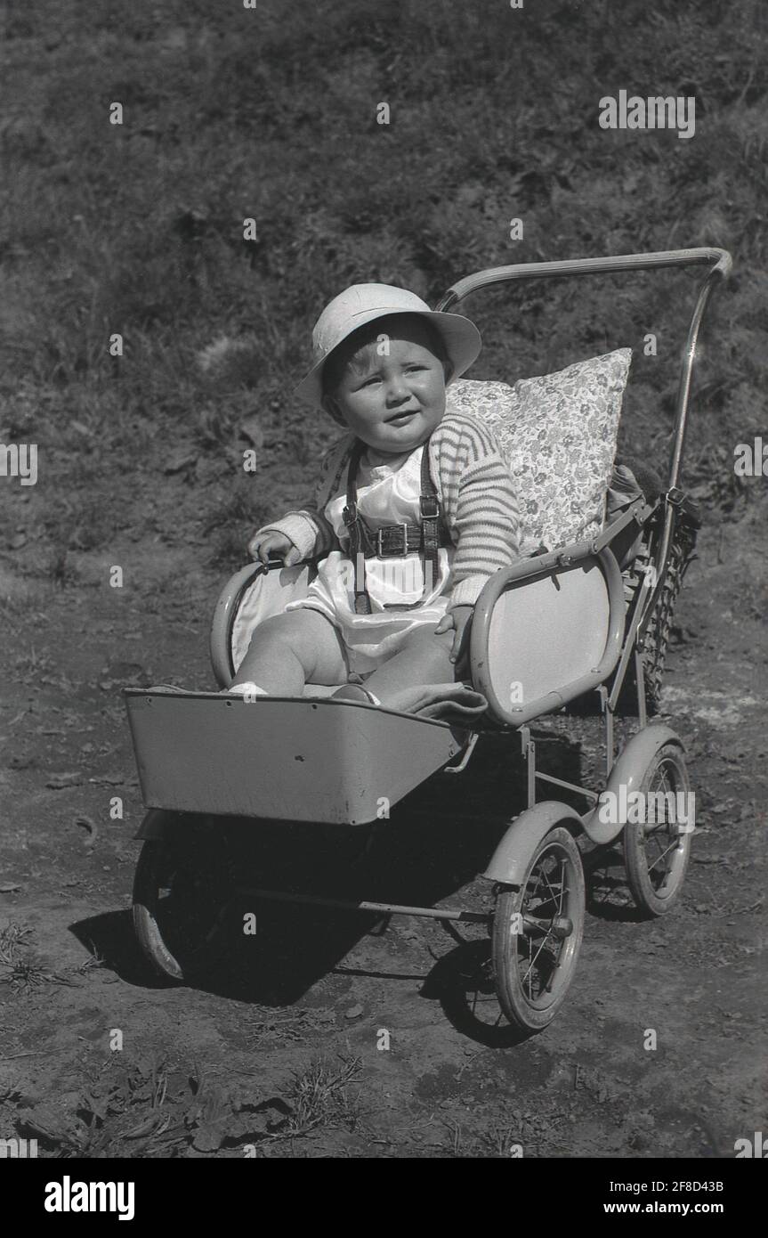 1950s, historical, outside on a summer's day, an infant child wearing a sunhat, sitting in a metal-frame pushchair or buggy of the era, with attached footrest and a pillow for back support, England, UK. Stock Photo
