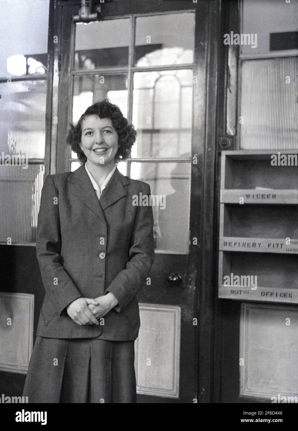 1950s, historical, a young female employee, a secretary or clerical worker, standing for a picture in her company office, with a wooden intray box for letters and correspondence fixed to the wall beside the glass-paneled door, England, UK. The office workplace in this era was formal one and employees were expected to dress in a certain way. Wearing a smart tailored wool suit, a jacket and skirt combination as she is, would present a professional image and would have been the appropriate clothing for a young female in a company office environment at the time, Stock Photo