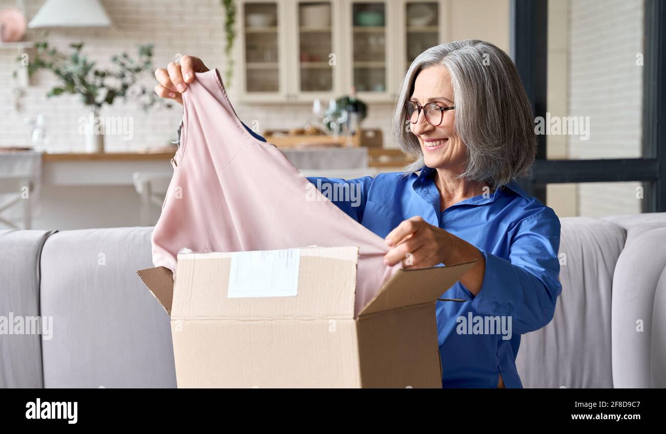 Senior mid age 60s woman opening parcel box at home on the couch. Stock Photo