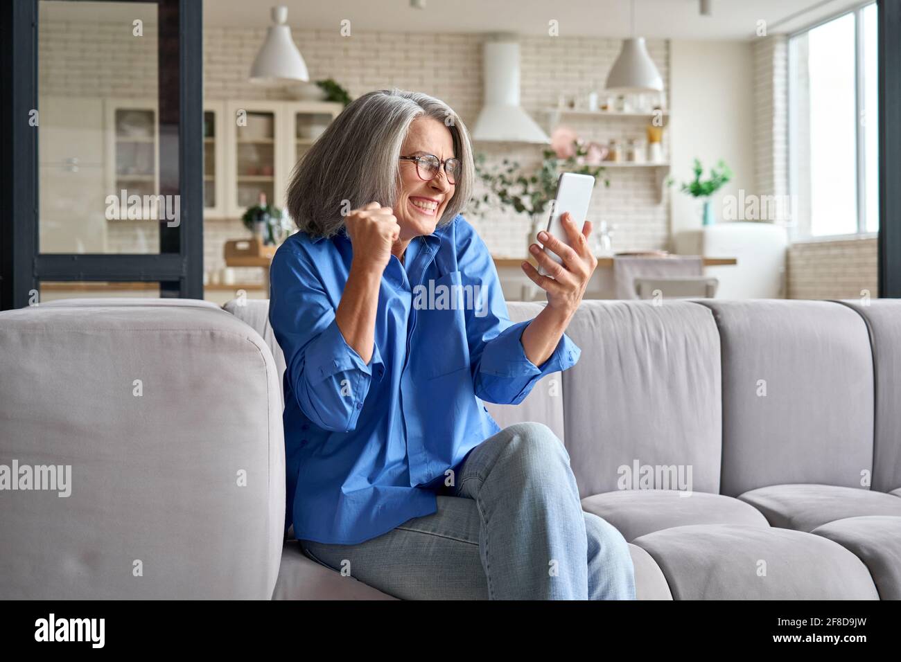 Surprised exited mature woman 60s aged at home with cellphone. Stock Photo