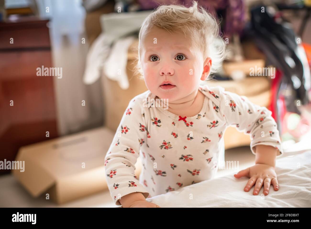 child with frightened eyes stands by bed. girl of 1 years learns to walk and is afraid to take a step Stock Photo