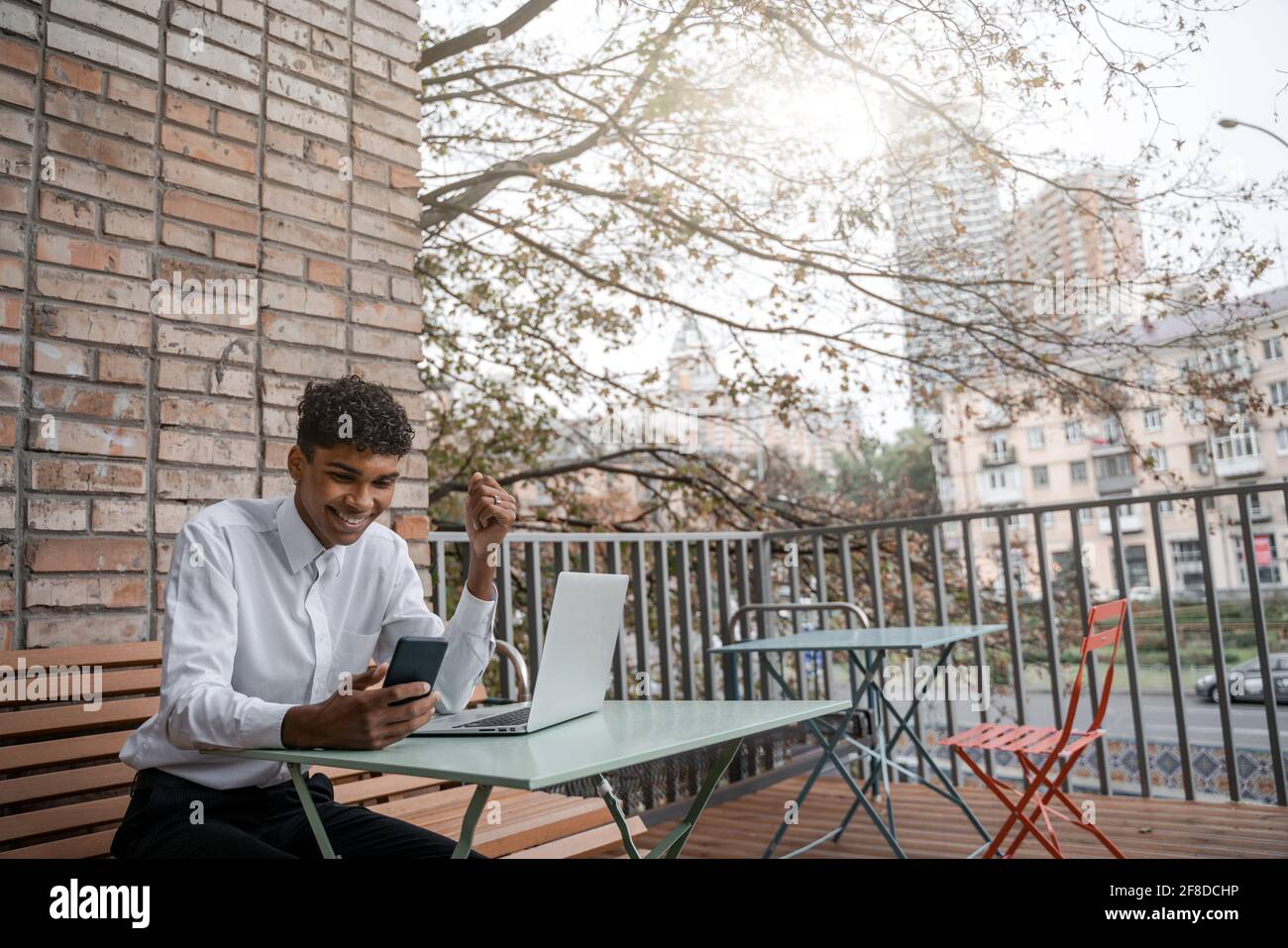 A black man sits on a summer terrace or veranda near a cafe. The guy works at the laptop. View of the spring business downtown Stock Photo