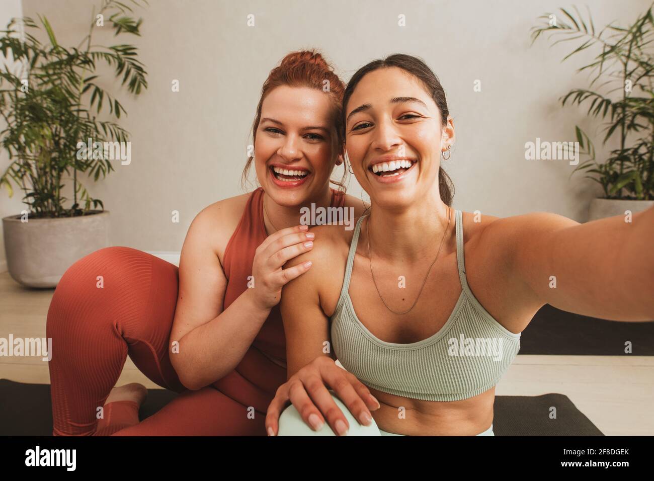 Cheerful female friends taking selife at yoga gym. Women in sports wear taking a break from workout and talking selfie at fitness studio. Stock Photo