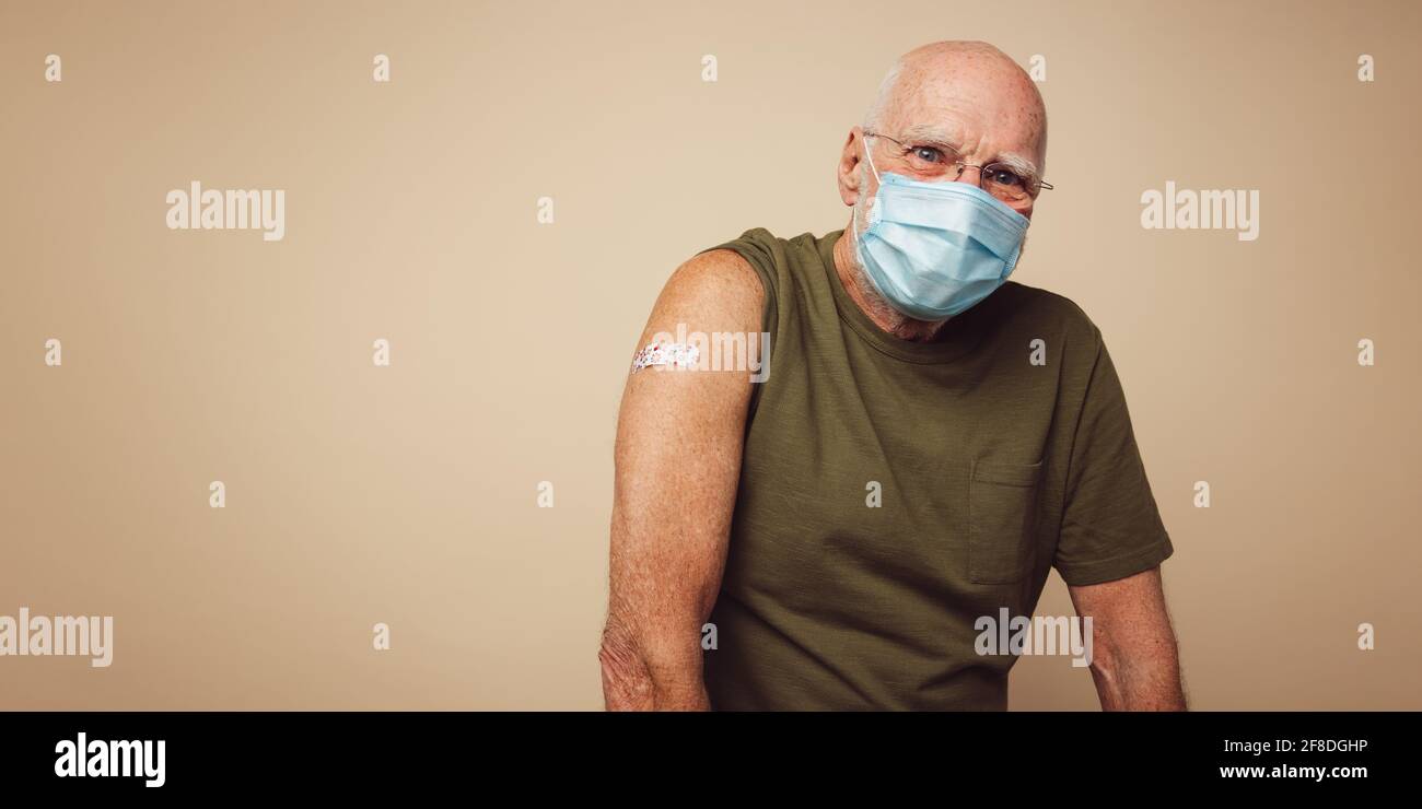 Portrait of senior man with protective face mask getting vaccine. Mature man against brown background after receiving corona virus vaccination. Stock Photo