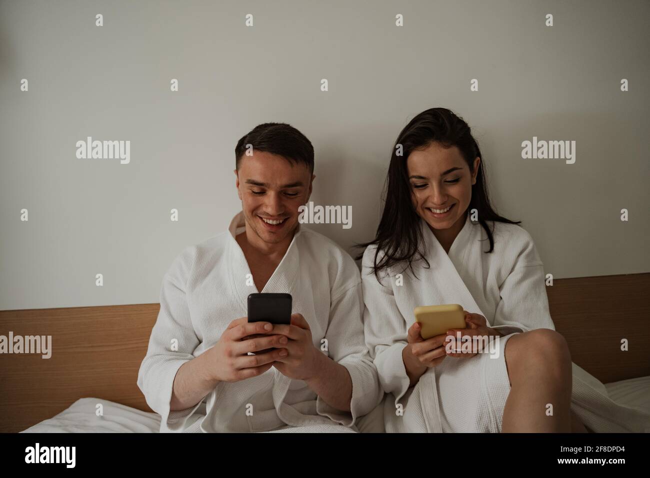 Morning of happy young couple in white bathrobes in bed at home, using phones Stock Photo