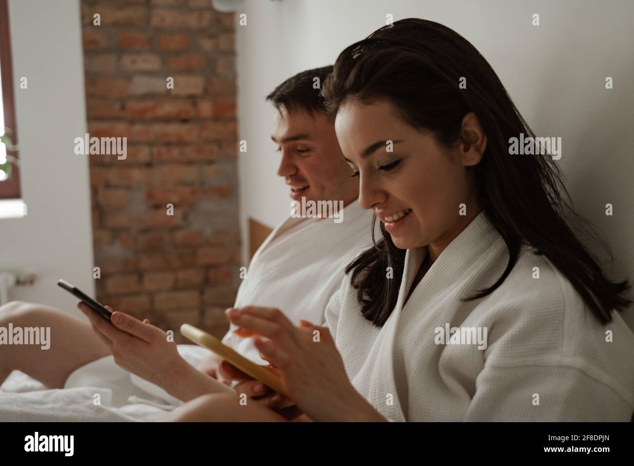 Morning of happy young couple in white bathrobes in bed at home, using phones Stock Photo