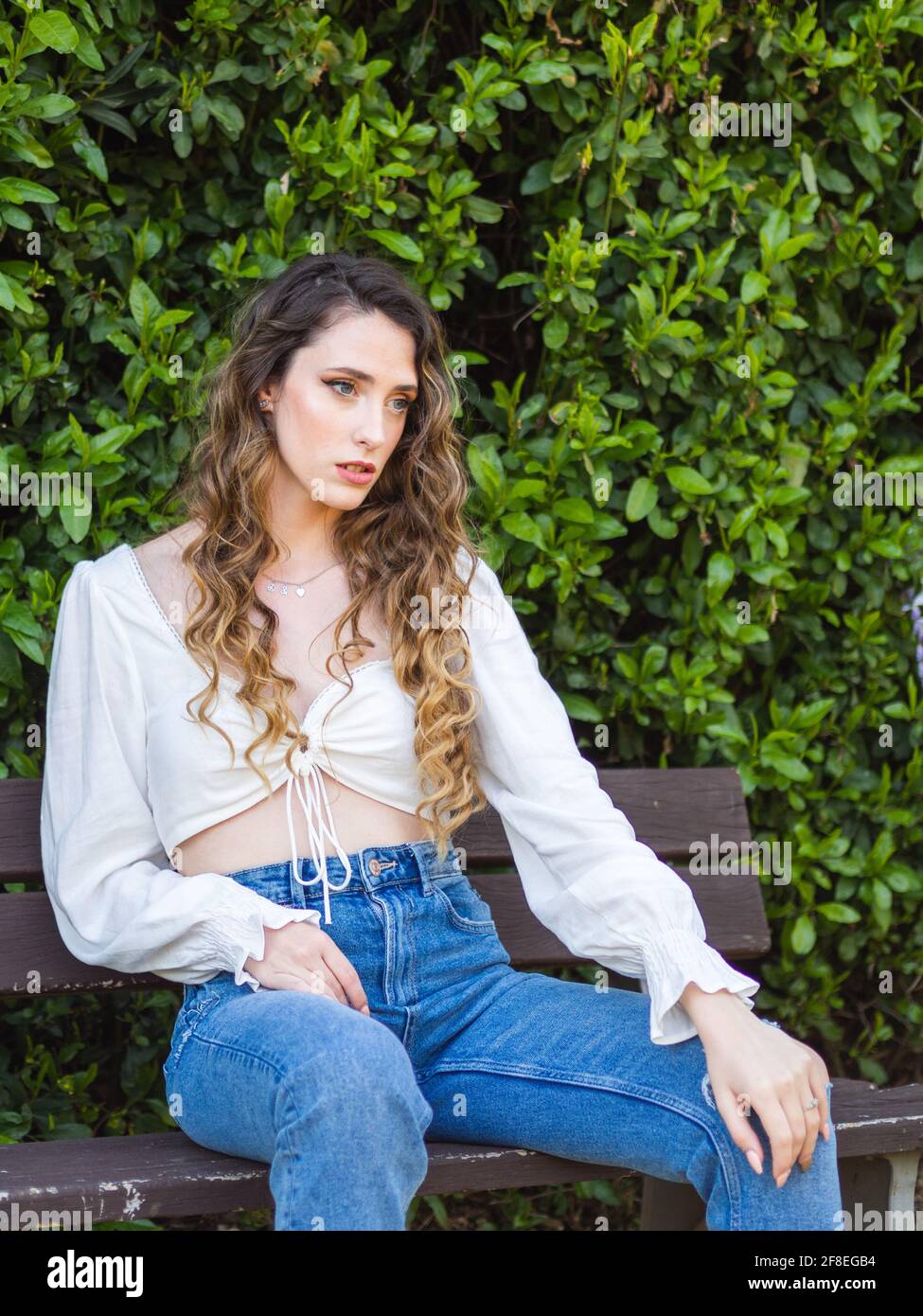 Young Spanish female wearing jeans a white blouse is sitting on a park springtime Stock Photo -