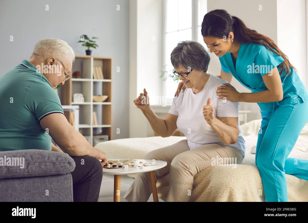 Supportive care giver watching couple of her happy senior patients play checkers Stock Photo