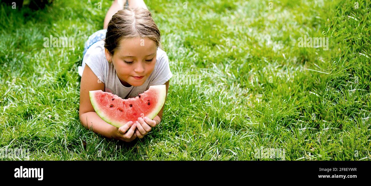 Portrait of a pretty girl 7-8 years old eating a piece of watermelon on the grass in the garden. Summertime. Staycation Stock Photo