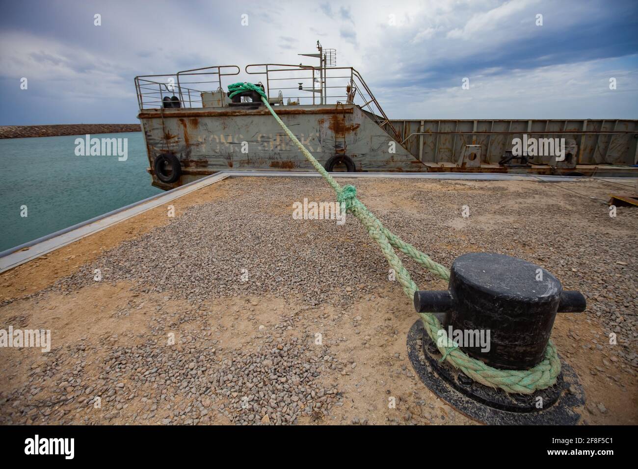 Aktau, Kazakhstan - May 19, 2012: Seaport and loading terminal. Cargo ship or barge in dock. Bollard with green rope on foreground. Cloudy sky. Stock Photo