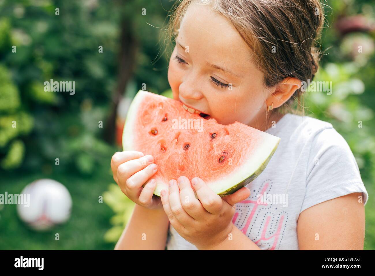 Portrait of a pretty girl 7-8 years old eating a piece of watermelon on the grass in the garden. Summer lifestyle Stock Photo