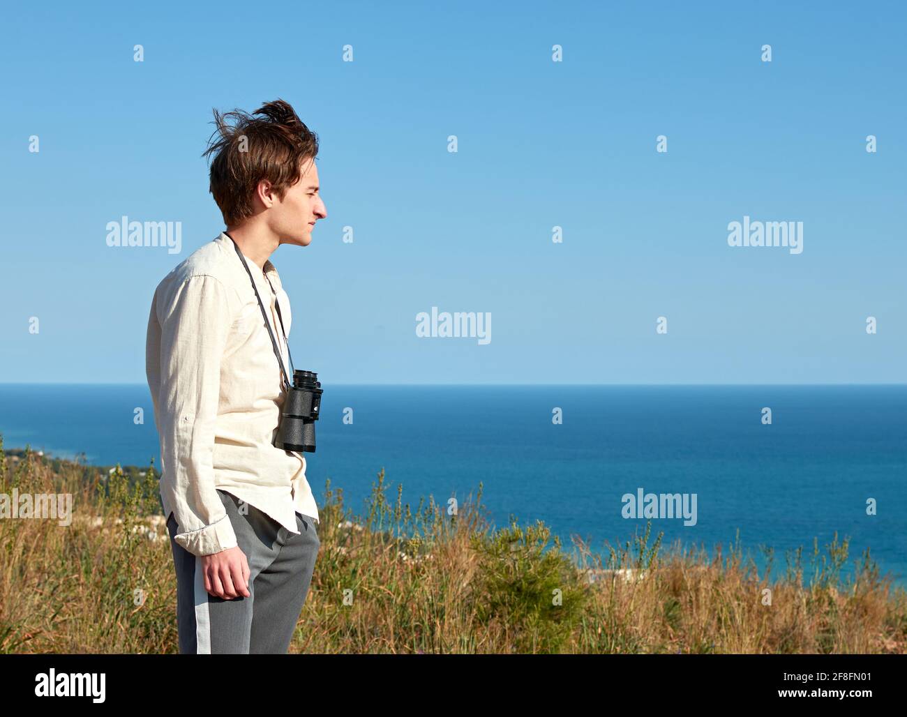 Side view of a young man with binoculars hanging around his neck in front of a sea on a windy day Stock Photo