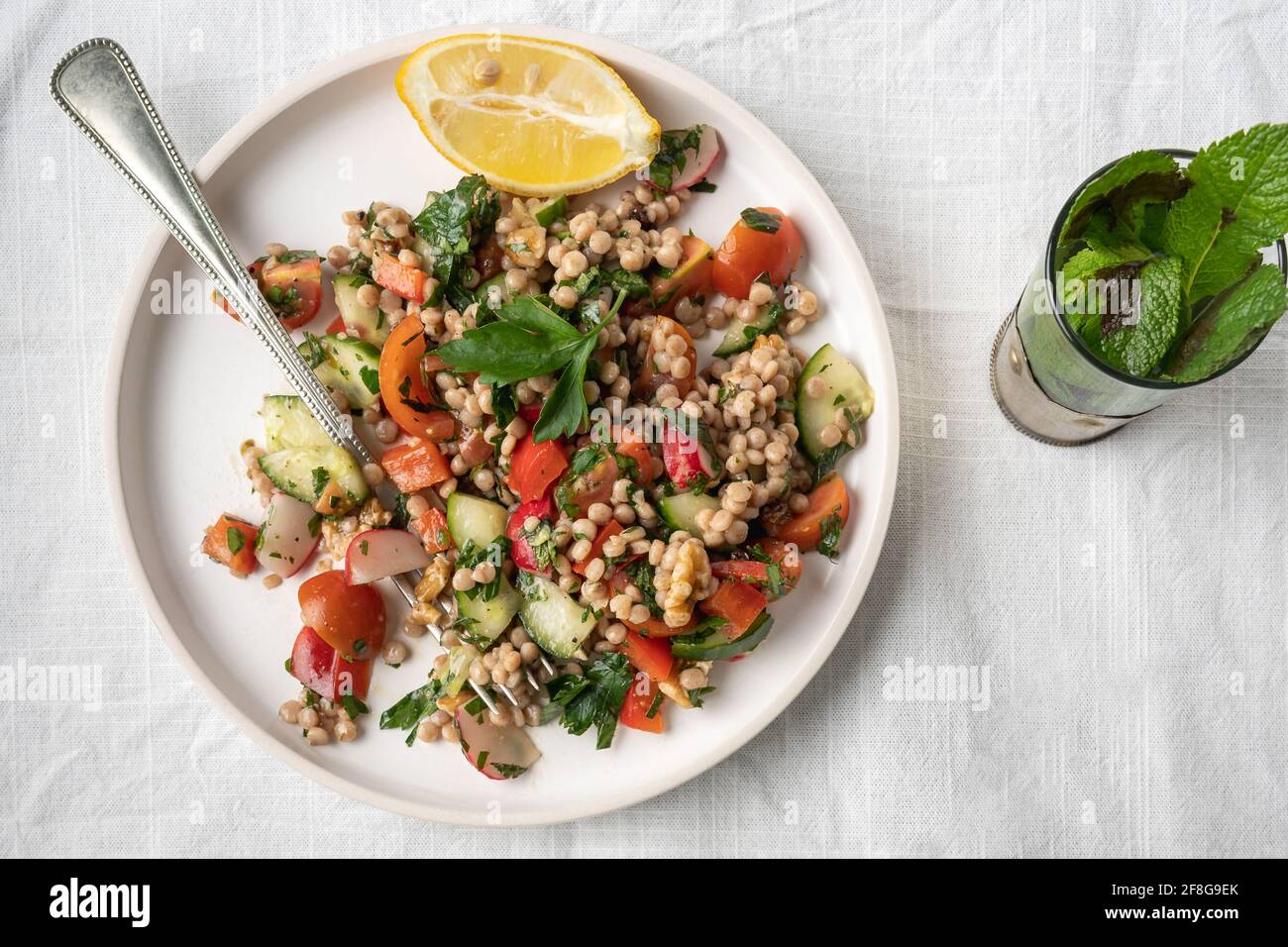 Flatlay of a single plate of a healthy vegan wholewheat pearl couscous salad with lemon and fresh vegetables on a white plate served with mint tea on Stock Photo