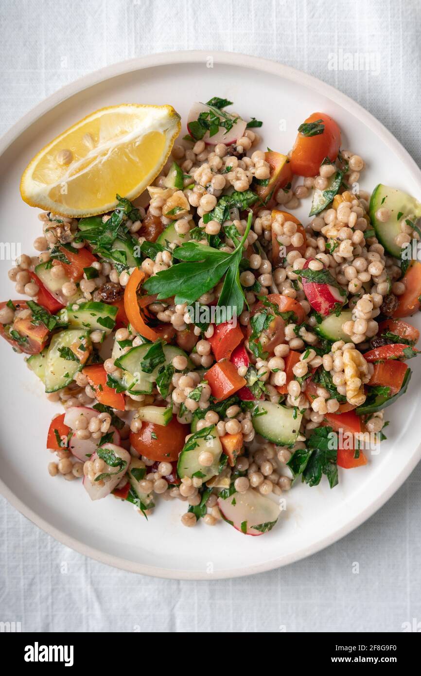 Close-up of a single plate of a healthy vegan wholewheat pearl couscous salad with lemon on white plate and white background, flatlay, vertical Stock Photo