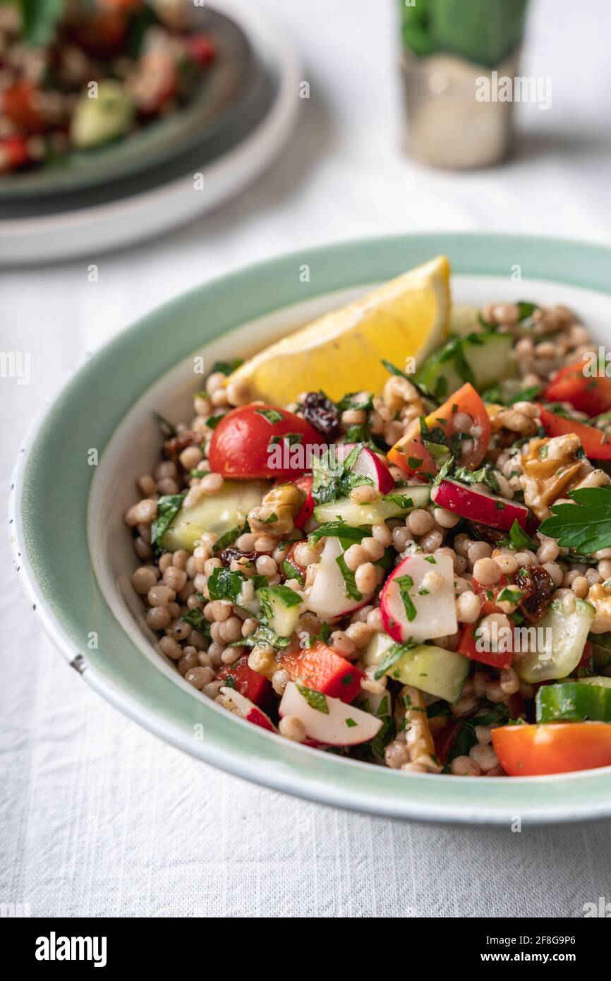 Close-up side view of two bowls with healthy vegan wholewheat pearl couscous salad with lemon ad fresh vegetables on a white background, with copy spa Stock Photo
