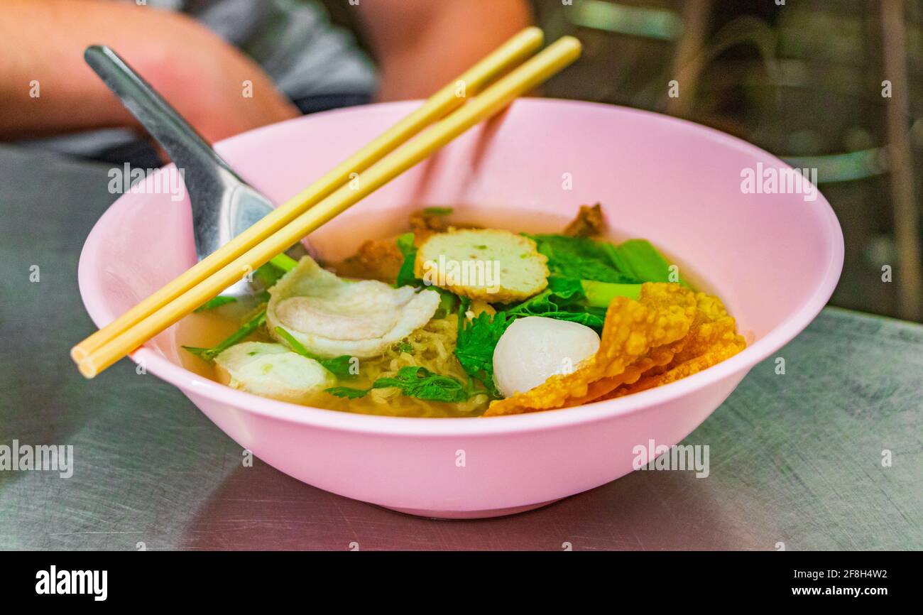 Thai noodle soup with fish balls in pink bowl with chopsticks and spoon