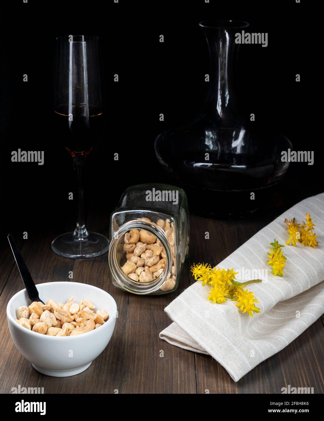 A white bowl containing peanuts and other dried fruit near glass containers with black wine and yellow flowers of sedum palmeri Stock Photo