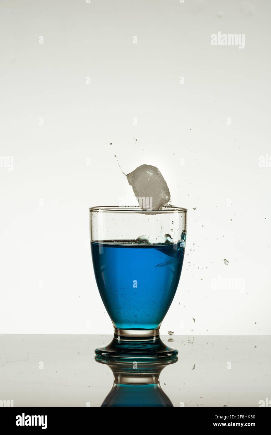 Ice cube falling into a glass of blue drink isolated on the reflective ...