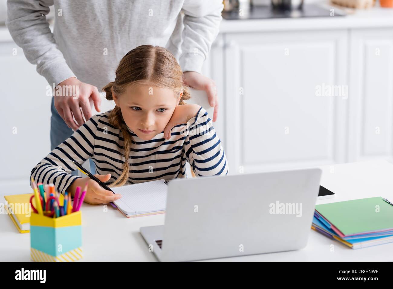 father standing behind schoolgirl looking at laptop during online lesson, blurred foreground Stock Photo