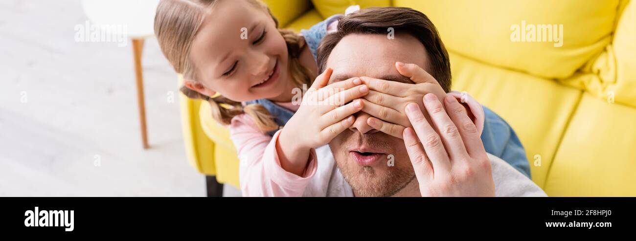 happy child covering eyes of surprised father while having fun at home, banner Stock Photo