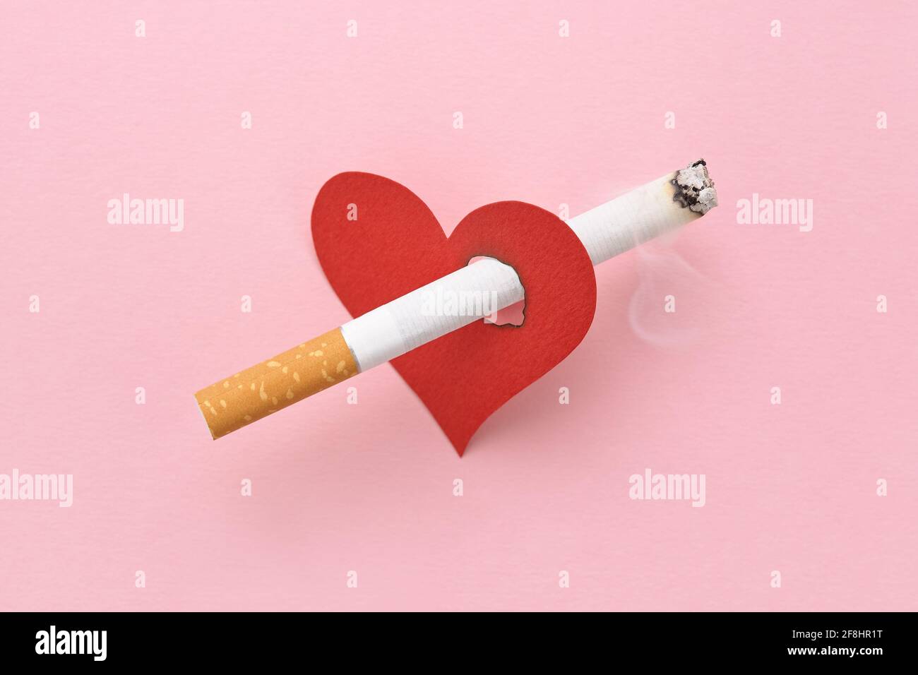 A red heart impaled by a burning cigarette, the harm of smoking, destroying health. Stock Photo