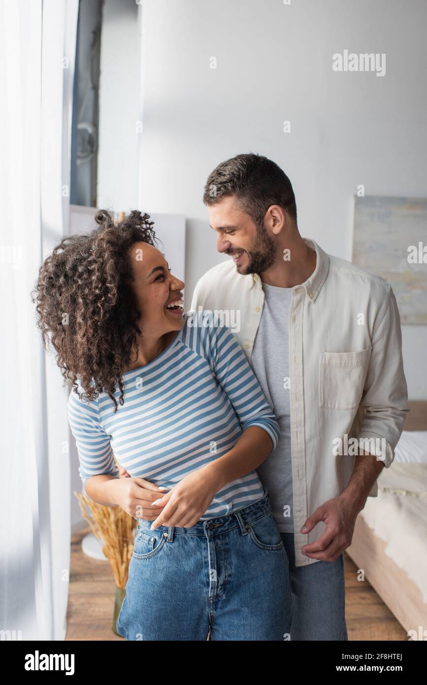 happy multiethnic couple laughing while hugging at home Stock Photo - Alamy