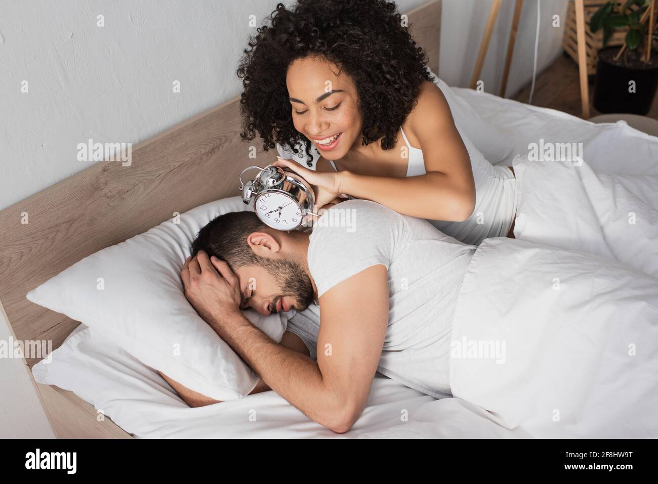 happy african american woman holding alarm clock near displeased man sleeping on bed Stock Photo