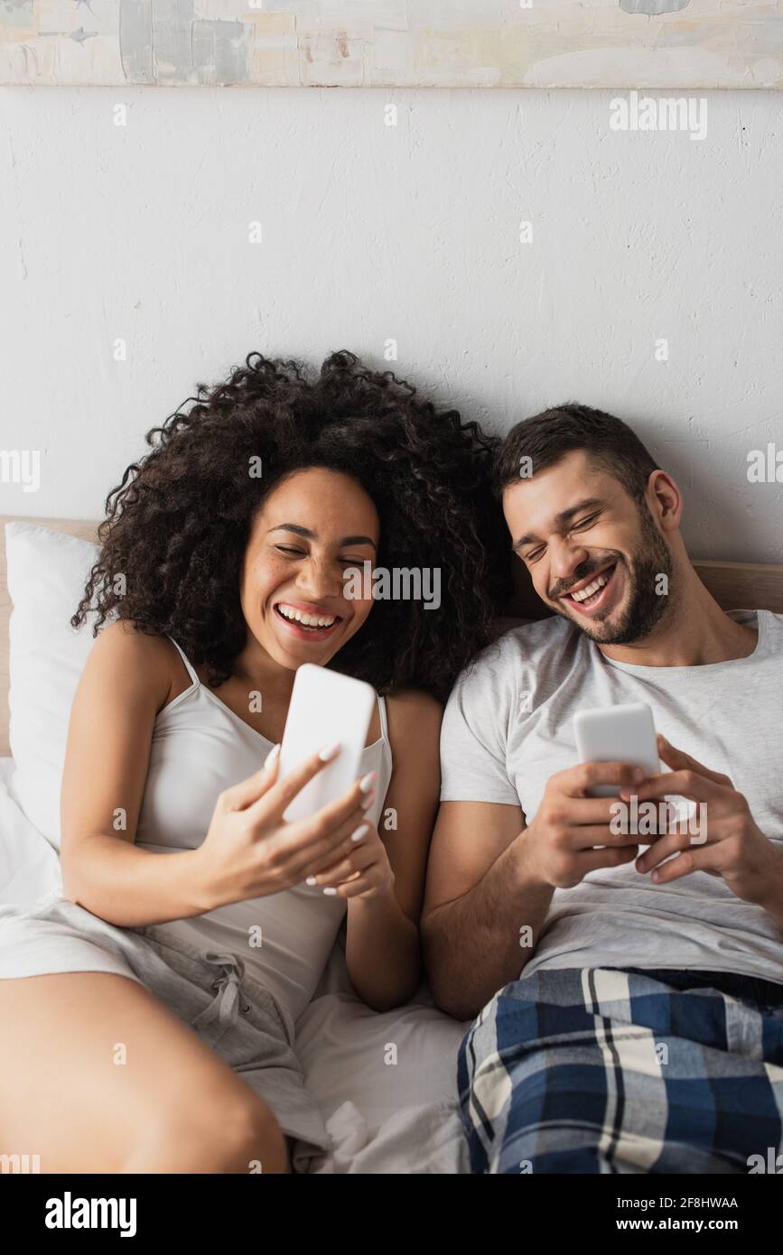 cheerful interracial couple using cellphones and laughing in bedroom Stock Photo
