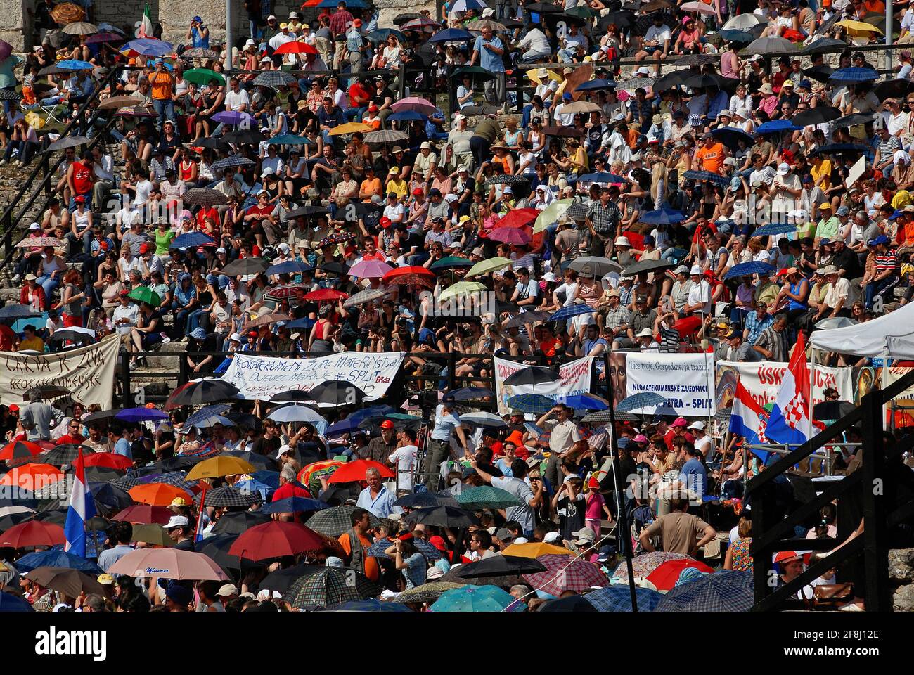 Parasols and umbrellas dot the ancient stone terraces of the Roman Arena at Pula, Istria County, Croatia, as the audience enjoying the music at a Roman Catholic festival and open-air service shelter from fierce June sun.  The amphitheatre, one of the six largest surviving ancient Roman arenas, once held up to 23,000 people, but the terraces can now seat only about 7,000.  Pula Arena is now a major music venue and has staged concerts by Foo Fighters, Luciano Pavarotti, Elton John, Sting, Leonard Cohen and David Gilmour. Stock Photo