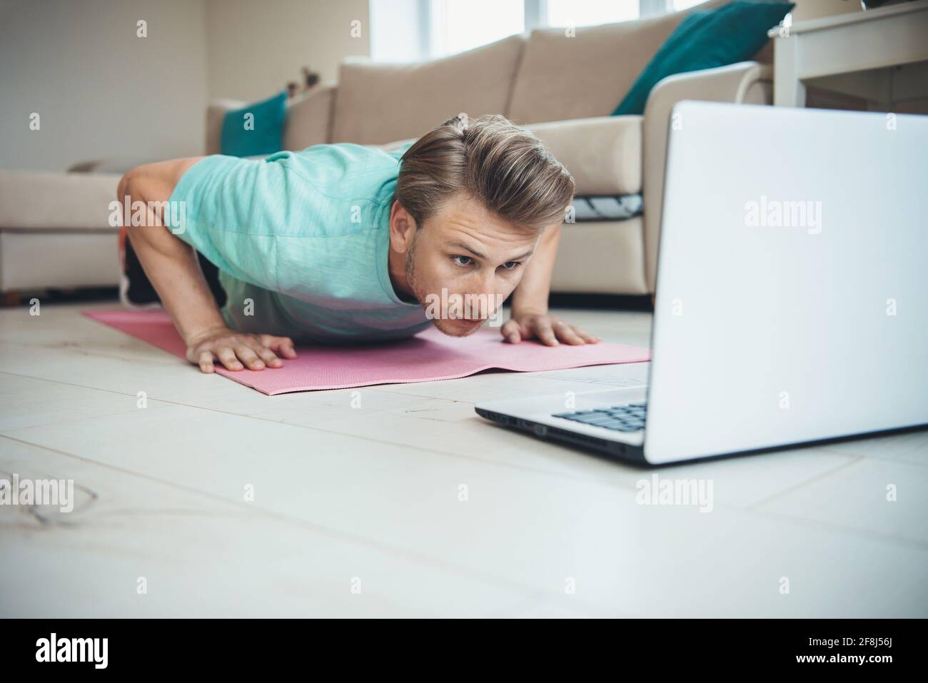 Caucasian man with blonde hair is doing push-ups on the floor while using a laptop Stock Photo