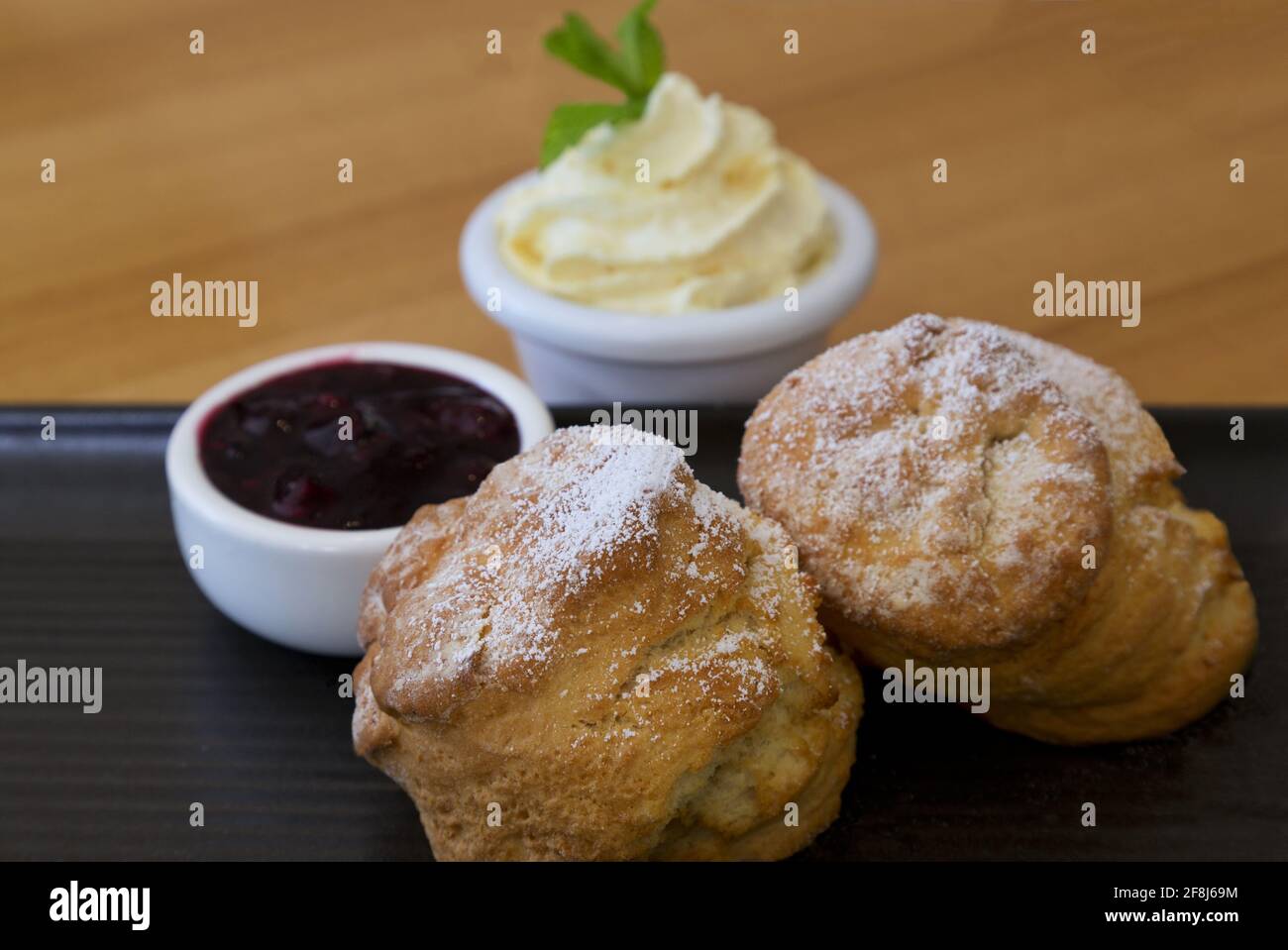 Hot scones sprinkled with powdered sugar and accompanied by whipped butter cream and red jam on rustic dark ceramic plate on light wood table. Stock Photo
