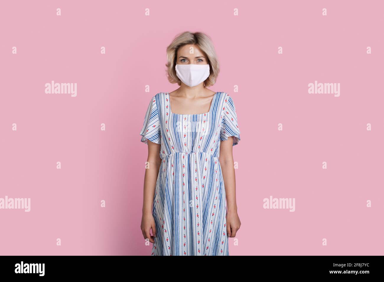 Blonde woman in a long summer dress is wearing a medical mask on a pink studio wall Stock Photo