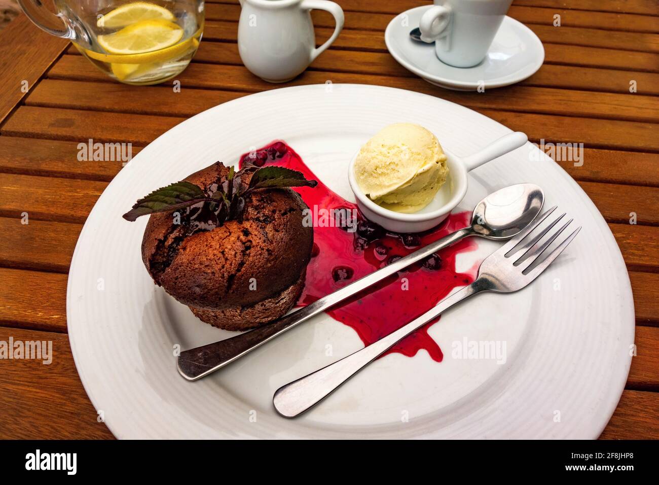 Chocolate fondant with vanilla ice and red forest fruit sauce on plate with cutlery, coffee, milk and glass of water on wooden table. Stock Photo