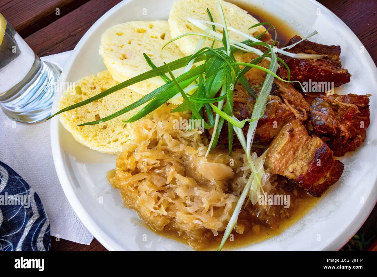 Roasted pork meat with stewed cabbage, dumplings and green onion on white plate, water,towel on wooden table, closeup. Czech republic tradition meal. Stock Photo