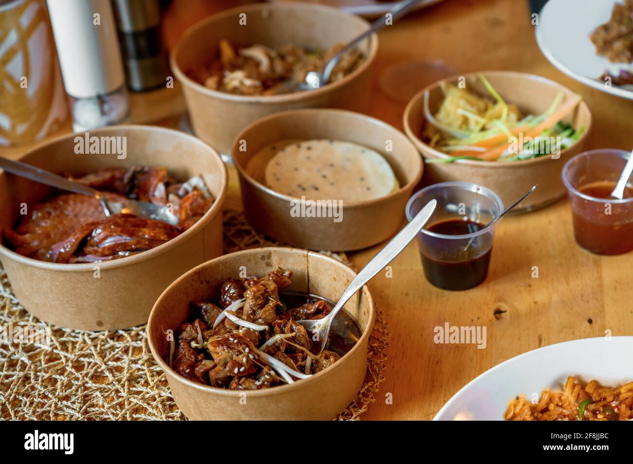 Table with many paper bowl with asian meal from restaurant delivery. Sliced baked duck meat, meat in spicy sauce, sesame pancake, vegetable salad . Stock Photo