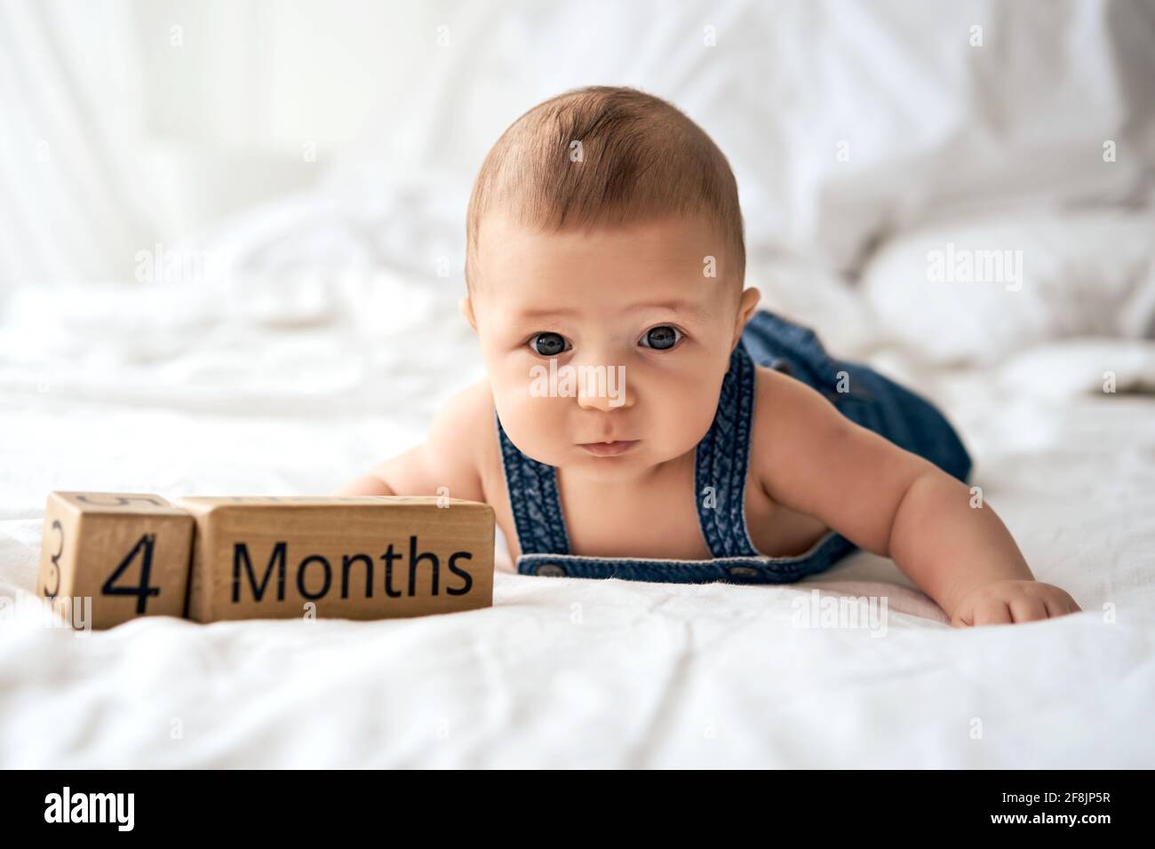 Adorable four month old baby boy in denim dungarees lying on white bed