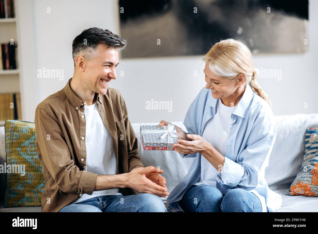 A middle aged married happy caucasian couple at home on the couch. A loving husband gives his beautiful wife a gift, small gift box in honor of a birthday, anniversary or March 8, they happy together Stock Photo