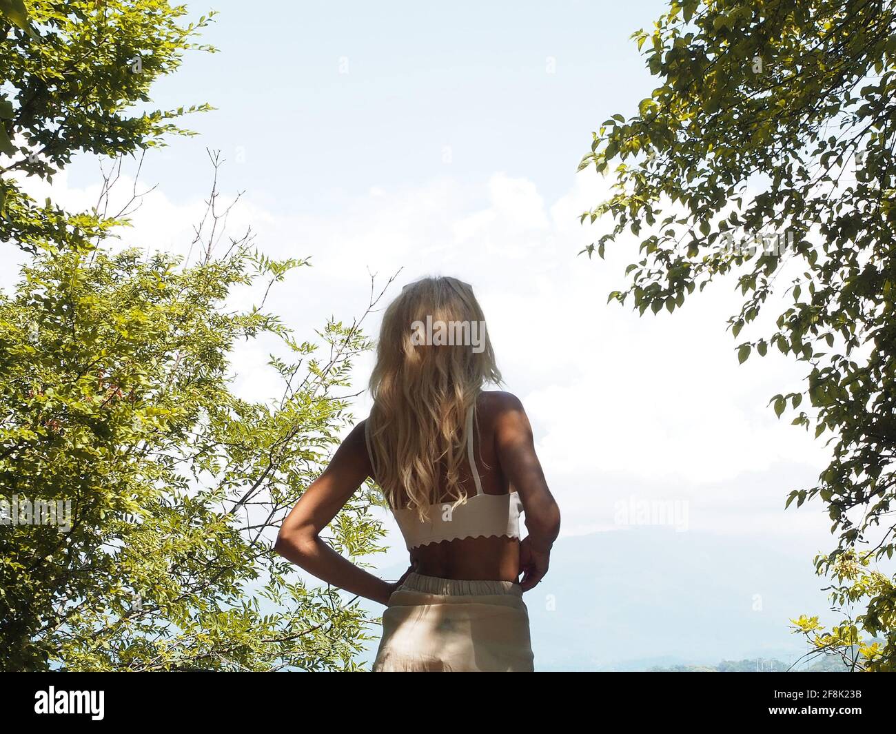A tanned blonde stands near green trees looking into light cloudy haze and the blue sky Stock Photo