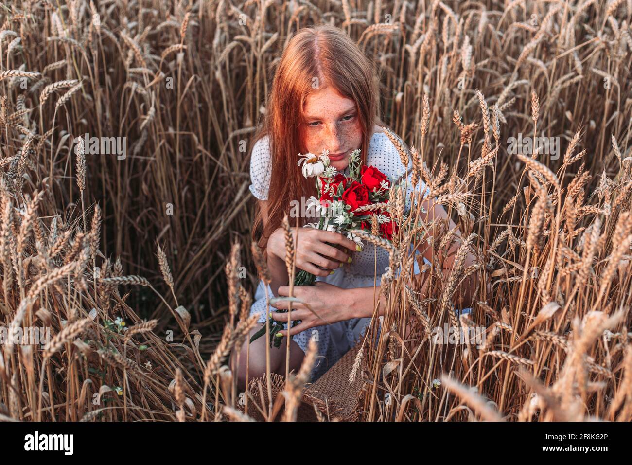 Portrait of young, teen, ginger girl with freckles in the wheat field with closed eyes, holding red bouquet. Copy space Stock Photo
