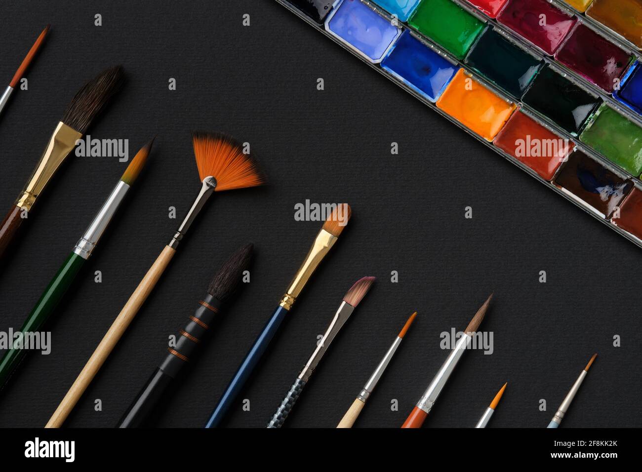 Different shape and size paintbrushes, set of watercolor paints on black paper sheet, top view, flat lay. Stock Photo