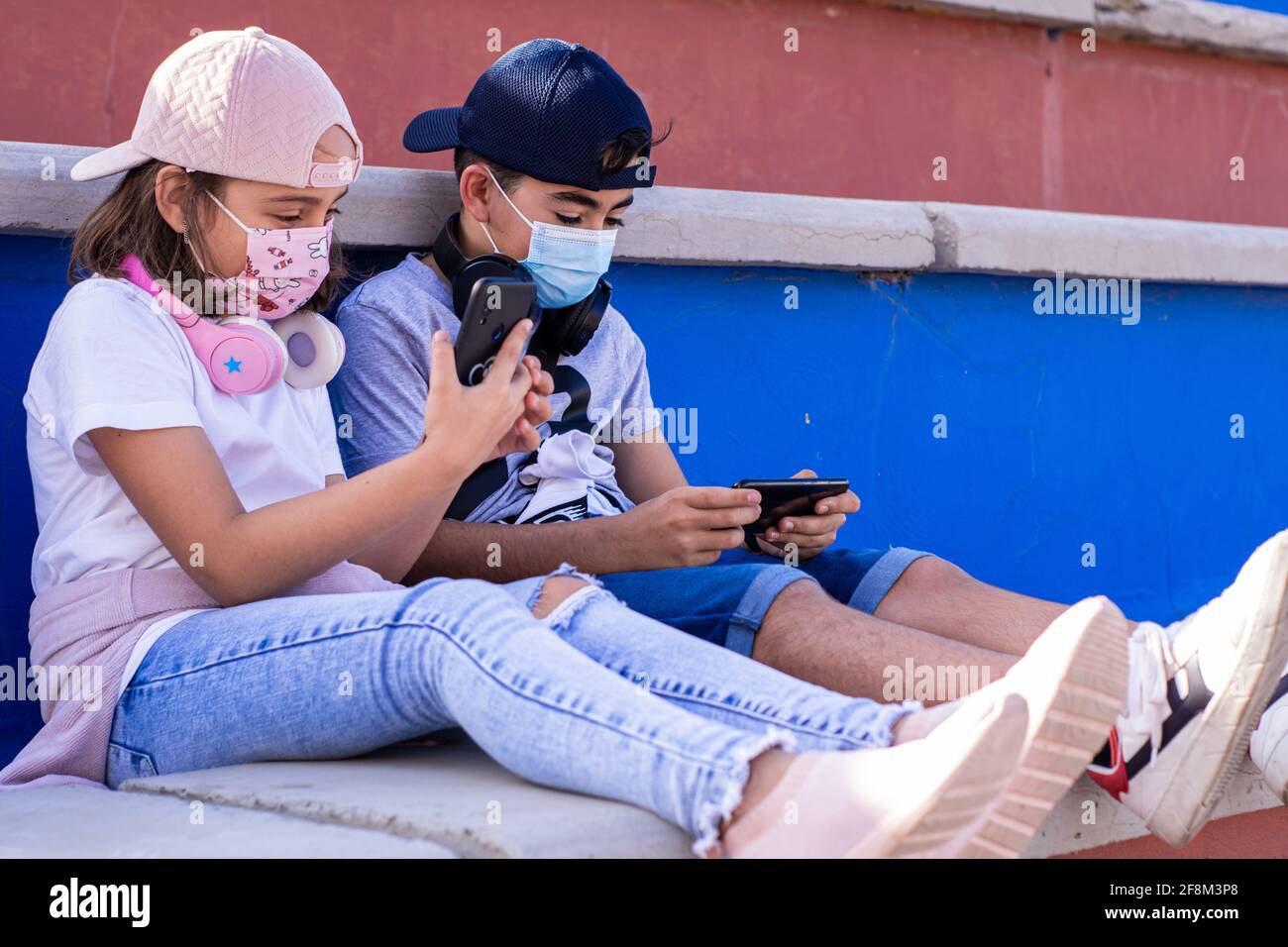 Two preteens, a boy and a girl, sitting on the bleachers with their masks on, use their mobile devices side by side. Stock Photo