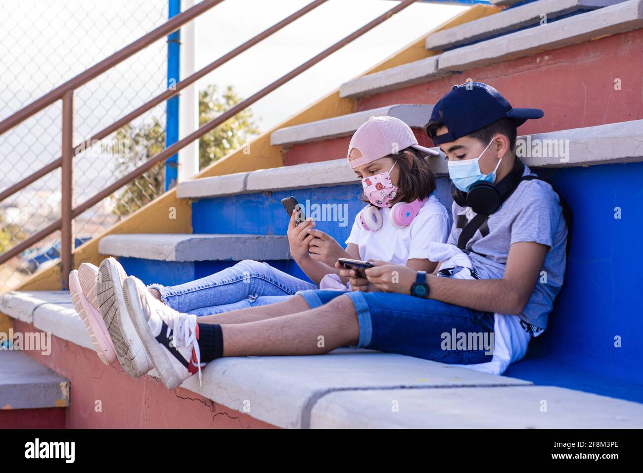 Two preteens, a boy and a girl, sitting on the bleachers with their masks on, use their mobile devices side by side. Stock Photo