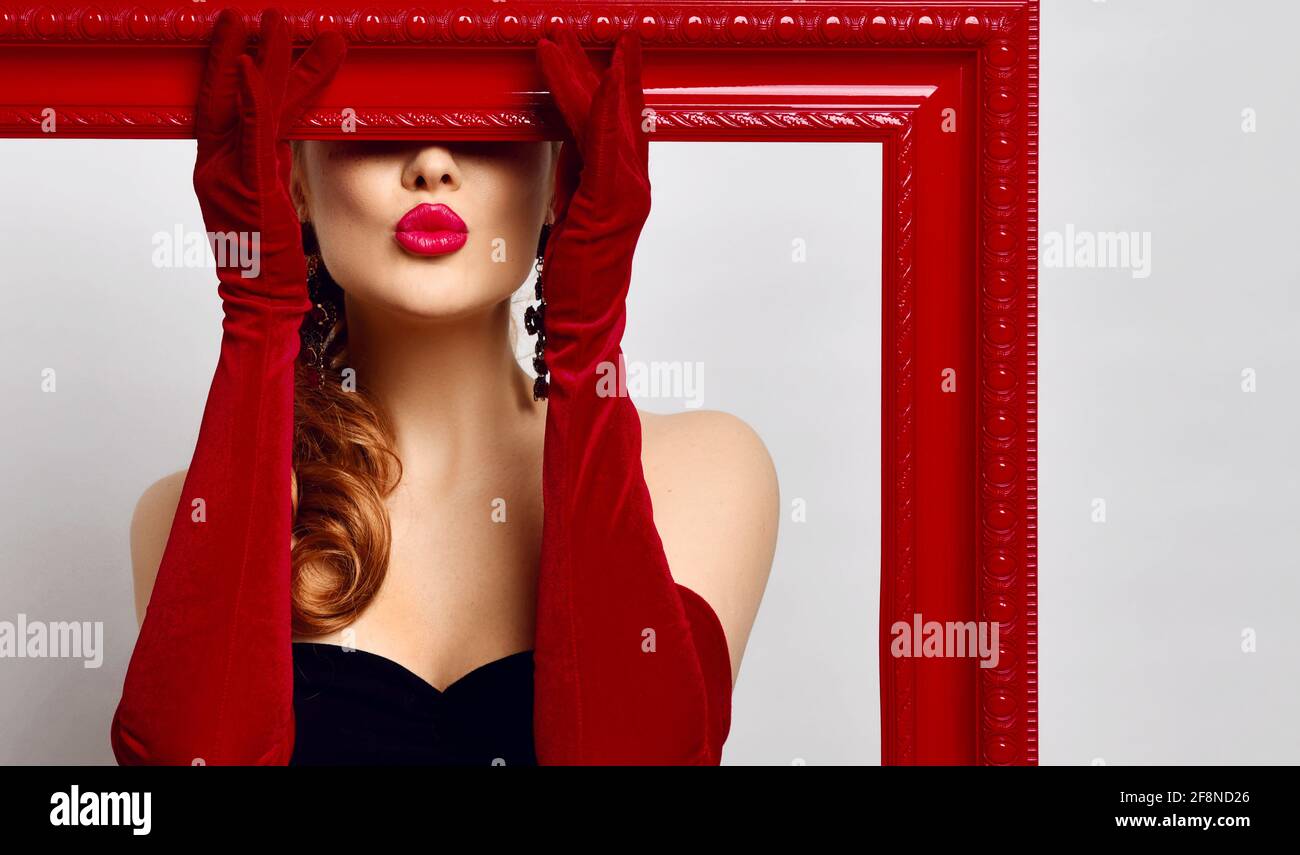 Young woman artist in black off-shoulder tight dress, red elbow-length gloves holds picture frame covering her eyes Stock Photo