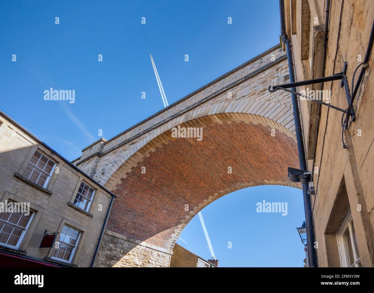 Jumbo jet aeroplane flying over railway bridge arch viaduct in blue sky with vapour trails. Low angle aircraft high in the sky looking up under old Stock Photo
