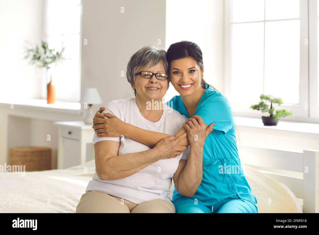 Portrait of happy caregiver together with senior patient sitting on bed in retirement home Stock Photo