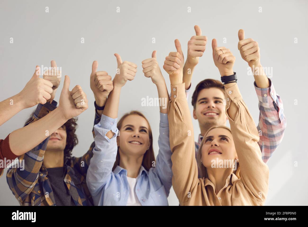 Group of smiling young people raising hands in air and giving thumbs-up all together Stock Photo