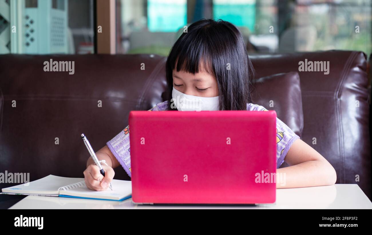 Asian child girl studying homework and wearing face mask during her online lesson at home for protect  Covid-19 Stock Photo