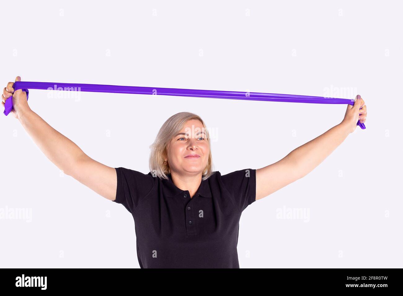 Fit senior blonde woman exercising with mauve resistance band against gym, smiling looking into distance on gray background with copy space. Stock Photo