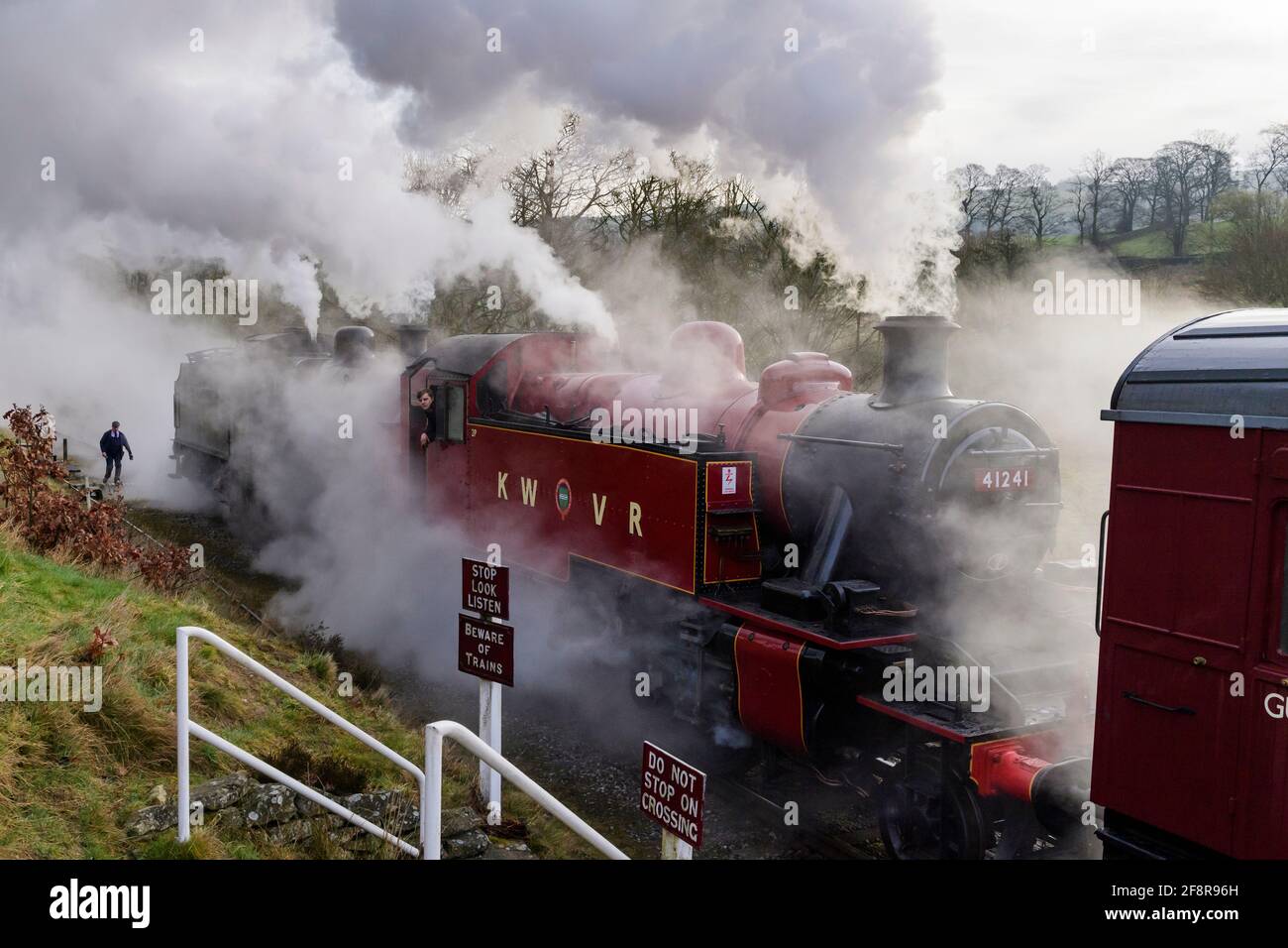 Historic steam trains (locos) puffing dramatic smoke clouds stopped at crossing (engine driver in cab) - heritage railway, KWVR, Yorkshire England UK. Stock Photo