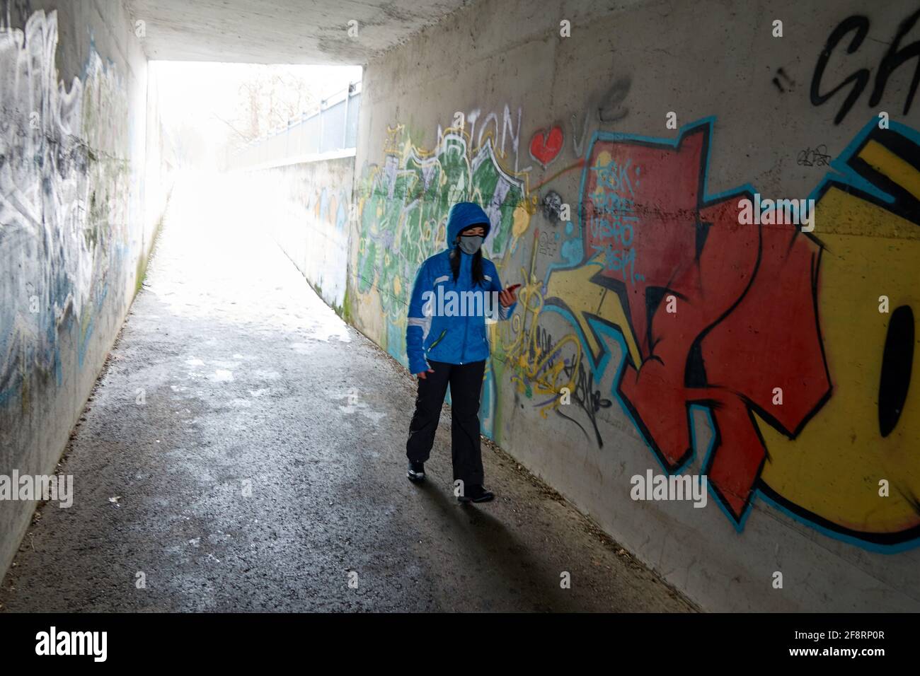 young woman in blue anorak with hood and protective mask runs through an underpass looking at her smartphone Stock Photo